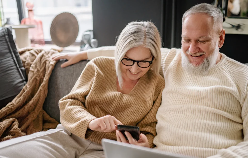 Older couple sitting together on a couch, smiling while looking at a smartphone with a laptop in front of them.